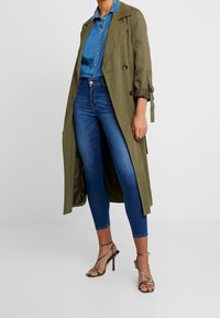 Trench coat verde oliva con diseño de doble botonadura, mangas enrolladas, usado sobre una camisa de denim y jeans ajustados azules, combinado con tacones de tiras.