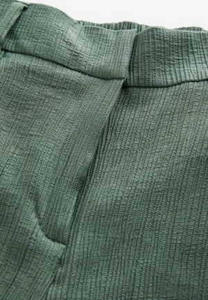 Zelená strukturovaná tkanina s zvrásněným povrchem, včetně širokého pásku a bočních kapes. Detailní pohled zdůrazňuje vzor materiálu.