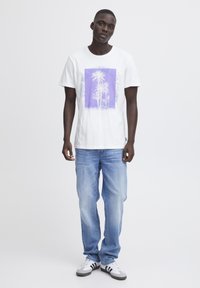 Camiseta blanca de algodón con un gráfico púrpura de palmeras y texto, combinada con jeans de color azul claro y zapatillas negras y blancas.