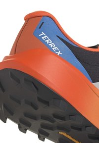 Nahaufnahme der Ferse eines orange-schwarzen Trailrunningschuhs mit blauem Etikett "TERREX" und robust profilierter schwarzer Sohle mit Continental-Logo.