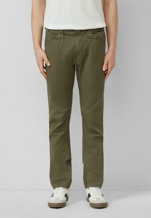 Homme portant un pantalon droit vert olive avec des poches et des baskets blanches, debout devant un fond gris clair uni.