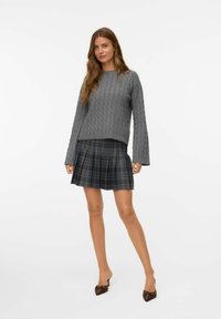Pull en maille gris avec des manches longues, associé à une jupe plissée à carreaux noir et gris. Le modèle porte des chaussures à talons marron.