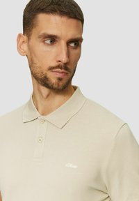 Beige Poloshirt aus strukturiertem Stoff, mit zweiknöpfigem Kragen und dezentem weißen Logo auf der Brust. Kurzarm und klassisches Kragen design.