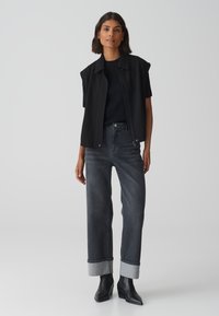 Zwarte mouwloze vest over een zwarte geribde top, gecombineerd met hoge donkere denim jeans met omgeslagen boorden en zwarte hakken.