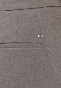 Szürke szövetnadrág, sima textúrával, hátsó zsebmintázattal és piros-fehér díszítéssel ellátott apró emblémával a zseben.