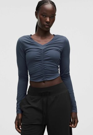 Vrouw met gevlochten haar die een geplooide, blauw crop top met lange mouwen en zwarte broek met trekkoord draagt, staand tegen een effen achtergrond.
