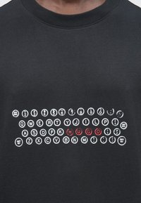 Juodas marškinėlis su baltais, rašomąja mašinėle stilizuotais klavišais išdėstytais eilėmis, o trečioje eilėje raudonomis raidėmis pabrėžtas užrašas „HUGO“.