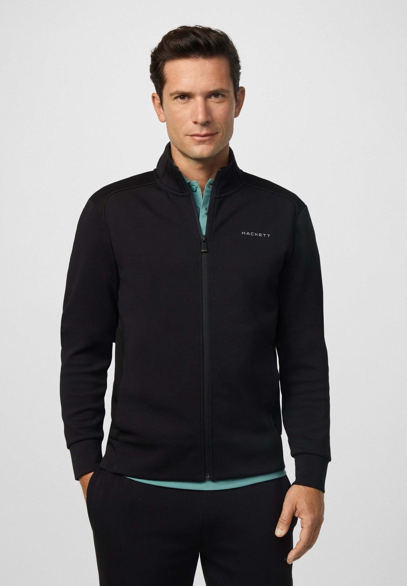 Muškarac u crnoj jakni na patent zatvarač preko zelene košulje s kragnom, stoji uz jednostavnu svijetlu pozadinu.