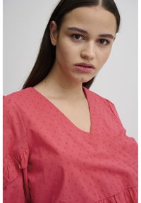Blouse en coton rouge avec un décolleté en V, des manches courtes à volants et un motif à pois texturé sur l'ensemble. Tissu lisse avec une coupe décontractée.