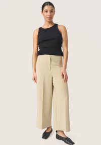 Sort ribbet tanktop og beige højttaljede, brede bukser. Flade, sorte slip-on sko med samlet detalje. Enkel, moderne design.