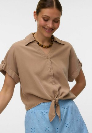 Frau lächelt, trägt ein kurzärmeliges, hellbraunes Hemd mit Knöpfen, das an der Taille gebunden ist, und einen blauen Rock mit Lochmuster, mit Perlenkette und kleinen Creolen.