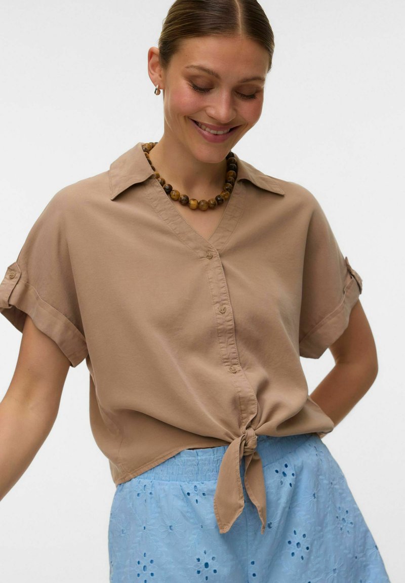 Femme souriante, portant une chemise beige à manches courtes boutonnée et nouée à la taille, ainsi qu'une jupe bleue à motifs œillets, avec un collier de perles et de petites boucles d’oreilles créoles.