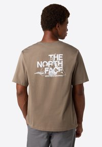 Mies, jolla on lyhyet kiharaiset hiukset, pukeutuu ruskeaan t-paitaan, jossa on valkoinen "THE NORTH FACE" -logo ja vuorireunat selässä.