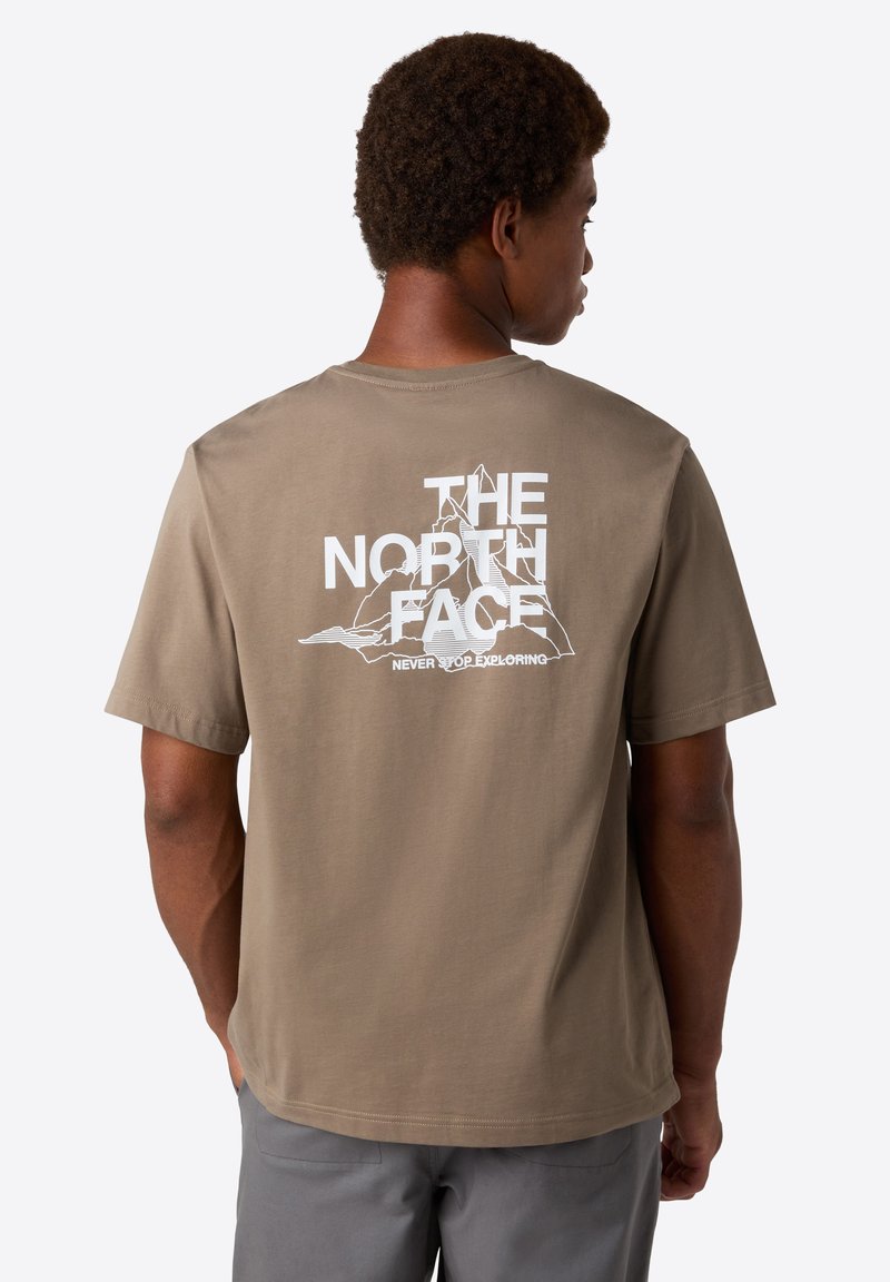 Mies, jolla on lyhyet kiharaiset hiukset, pukeutuu ruskeaan t-paitaan, jossa on valkoinen "THE NORTH FACE" -logo ja vuorireunat selässä.