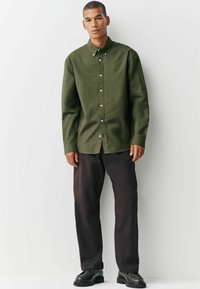 Chemise boutonnée vert olive avec un col classique, une poche avant et des boutons blancs. Assortie à un pantalon foncé et des chaussures noires.