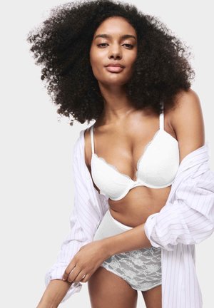 Reggiseno in pizzo bianco con bordi scallop, abbinato a un perizoma in pizzo a vita alta. Il modello indossa una camicia a righe larga.