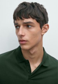 Donkergroene polo met een gestructureerde stof, voorzien van een kraag en twee knopen, gedragen door een model met kort, warrig haar.