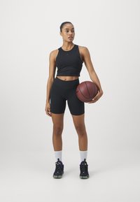 Camiseta sin mangas atlética negra y pantalones cortos hasta la rodilla. Zapatillas negras con acentos morados, sosteniendo un balón de baloncesto marrón. Fondo simple.