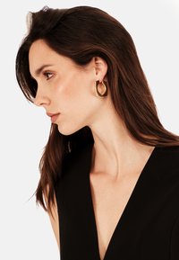 Zlaté geometrické náušnice se sochařsk�ým designem, který má duté středy a lesklý povrch, nosí modelka s tmavými vlasy.