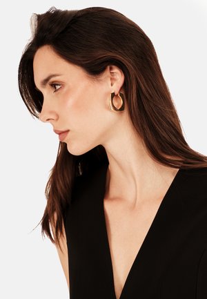 Złote geometryczne kolczyki o rzeźbiarskim wzornictwie, z pustym środkiem i polerowanym wykończeniem, noszone przez modelkę z ciemnymi włosami.
