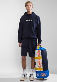 Sudadera azul marino con logo, pantalones cortos a juego y zapatillas blancas. Sosteniendo una bolsa de viaje colorida con gráficos y acentos amarillos.
