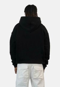 Crna dukserica s kapuljačom, opuštenog kroja, s velikom kapuljačom i dugim rukavima, u kombinaciji s bijelim hlačama s dva stražnja džepa.