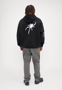 Felpa nera con una grafica di ragno bianca sul retro e un design di ragnatela sulla manica sinistra; abbinata a pantaloni cargo grigi e sneaker nere.