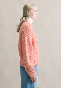 Gestrickter Pullover in leuchtendem Korallrot mit strukturiertem Muster, oversize Passform und geripptem Saum, kombiniert mit hellblauen Jeans.