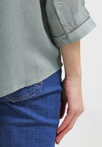 Blusa verde chiaro con texture e maniche arrotolate, con dettaglio dei bottoni. Abbinata a jeans blu con design della tasca posteriore.