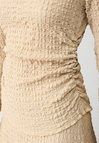 Top beige texturizzato con dettagli arricciati lungo i lati, realizzato in un tessuto leggero con una superficie increspata.