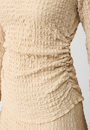Haut texturé beige avec des détails froncés sur les côtés, fabriqué dans un tissu léger à la surface plissée.