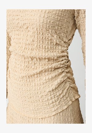 Beige textuur top met gathered details langs de zijkanten, gemaakt van een lichte stof met een gekreukeld oppervlak.