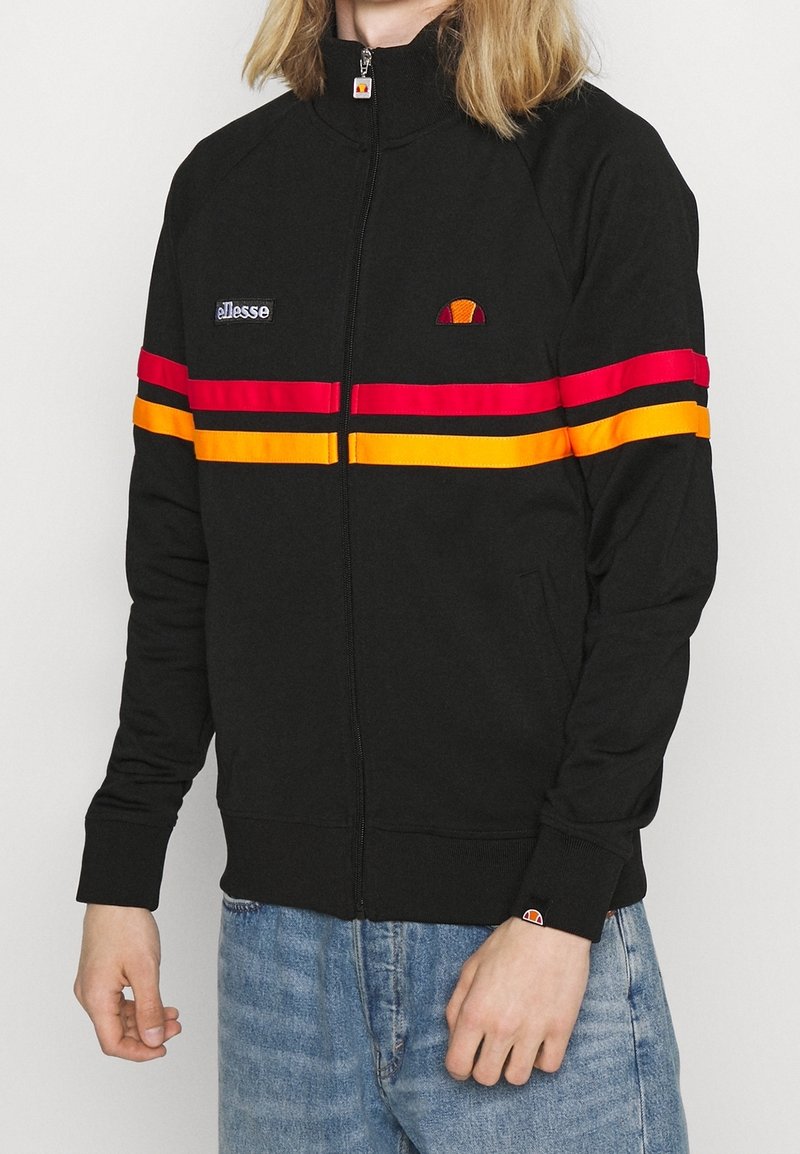 Personne portant une veste noire zippée avec des bandes horizontales rouges et orange et des logos sur la poitrine et les poignets, associée à un jean bleu.