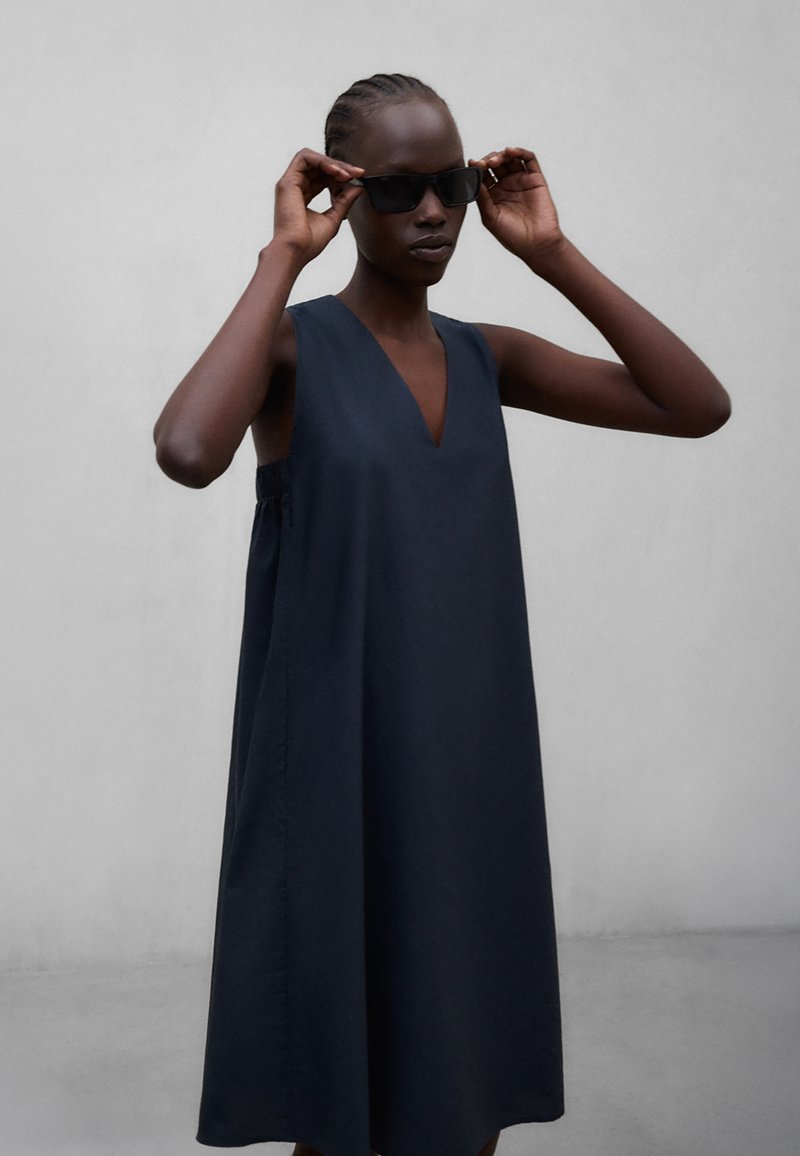 Femme portant une robe sans manches bleu marine à encolure en V, ajustant des lunettes de soleil rectangulaires noires devant un fond uni gris clair.