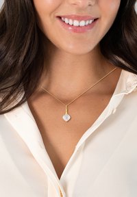 Guldkæde med en klar, facetteret, hjerteformet vedhæng. Designet har en glat, reflekterende tekstur. Båret med en cremefarvet bluse.