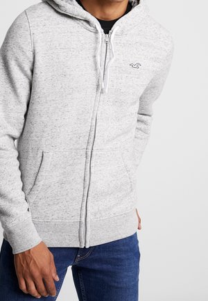 Mand iført lys grå lynlåshoodie med snore og kængurulommer, kombineret med blå jeans, stående mod en ensfarvet baggrund.