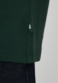 Ourlet en tissu vert foncé avec une petite étiquette attachée, porté sur un jean en denim bleu foncé.