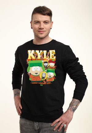 Mężczyzna w czarnej bluzie z postacią Kyle z South Park i tekstem "KYLE I learned something today" na przodzie, stoi z rękami na biodrach.