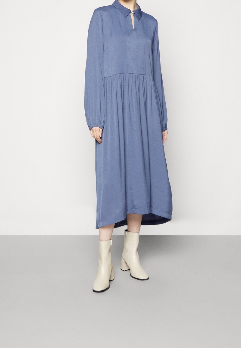 Femme portant une robe midi bleue à manches longues avec un col et une encolure goutte d'eau, associée à des bottines crème sur un fond uni.