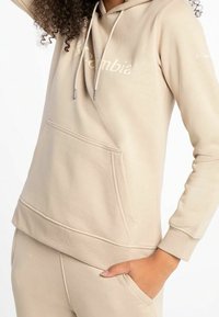 Sudadera beige con un bolsillo frontal, cordones ajustables y una textura suave. Presenta un logo sutil en el pecho y puños acanalados.