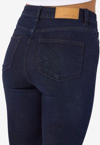 Gros plan d'une personne portant un jean denim bleu foncé avec des poches arrière et un patch en cuir beige portant l’inscription "NOISY MAY" sur la ceinture.