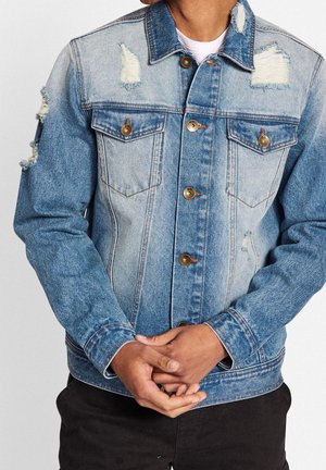 Mand iført lyseblå slidt denimjakke med metal knapper, stående med hænderne foldet foran sig, mod en ensfarvet baggrund.
