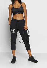 Schwarzer Sport-BH mit strukturiertem Muster und Logo, kombiniert mit schwarzen Jogginghosen, die auf einer Seite ein Logo tragen. Hält ein Springseil.