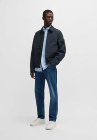 Giacca blu scuro con design a colletto, indossata sopra una camicia azzurra chiaro. Abbinata a jeans scuri e sneakers bianche. Tessuto morbido.