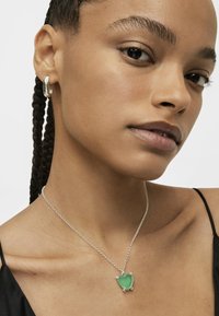 Collana in argento con un pendente a forma di cuore verde traslucido con accenti metallici, indossata su un collo nudo su uno sfondo neutro.