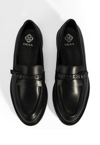 Zwarte leren loafers hebben een band met een gesp en decoratieve studs, een gladde textuur, een ronde neus en een geribbelde rubberen zool.