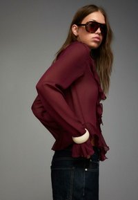 Burgundy chiffon blouse with ruffled trim and long sleeves, paired with dark denim jeans. Accessorized with a large white bracelet.