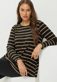 Top de rayas negras y beige de manga larga con escote redondo y textura acanalada, combinado con pantalones oscuros. Modelo sentado contra una superficie blanca.