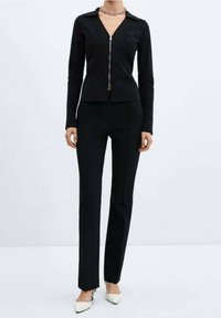 Black fitted jacket with a zipper, long sleeves, and a collar, paired with matching black tailored pants. Model wears white heeled shoes.