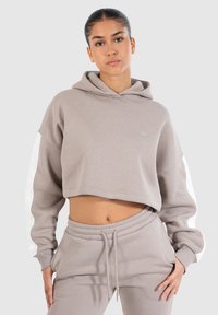 Korte beige hoodie gemaakt van zachte stof met witte accenten op de mouwen. Voorzien van een trekkoordcapuchon en een relaxte fit. Gecombineerd met bijpassende broek.